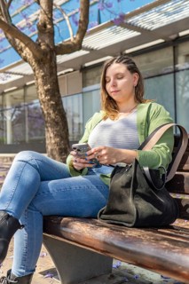 Young woman with braided hair, green jacket, and black bag using her smartphone on a sunny day. She