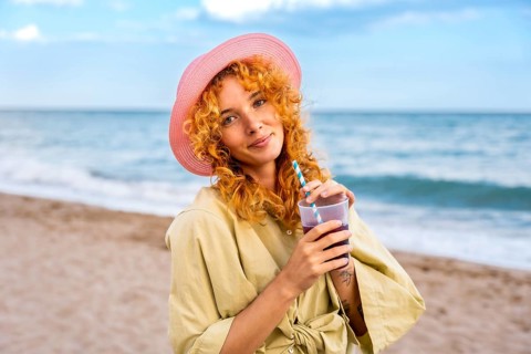 Curly haired woman wearing a pink straw hat enjoying a refreshing drink on the beach during a