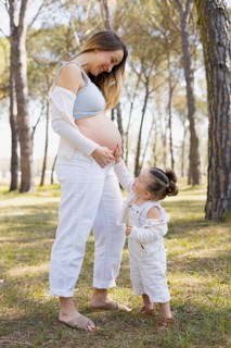 A pregnant woman stands in a serene forest scene with her young daughter, who gently touches her
