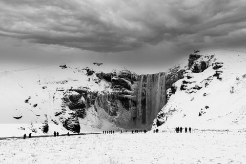 Tourists at Skogafoss waterfall, snowy and icy rock face, dark clouds above, black and white photo,