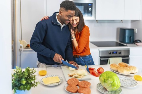 Young multiethnic couple hugging and slicing onions, preparing burgers for dinner in a modern white