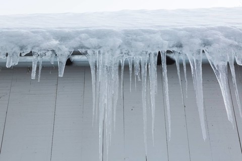 Long icicles and thick heavy ice hanging from roof edge of grey wood plank cladded building in
