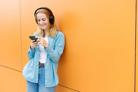 Smiling woman leans against a vibrant orange wall in Warsaw, listening to music on wireless