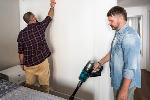 A gay couple is seen doing household chores together. One is vacuuming while the other wipes the