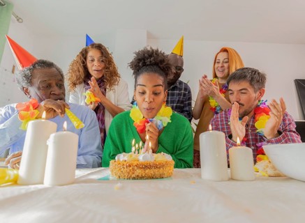 Group of friends celebrating a birthday with a cake and lit candles. Everyone is wearing party hats