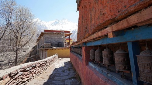 Prayer wheels along a brick wall with mountains and buildings in the background, Manang, trekking