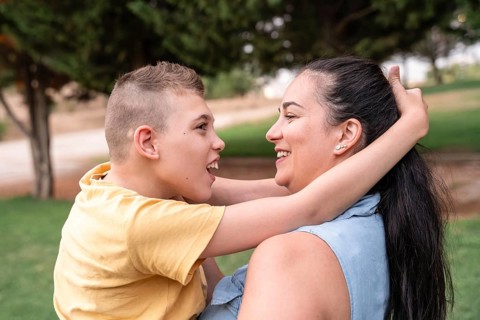A mother embraces her son with cerebral palsy outdoors. They share a joyful moment, highlighting