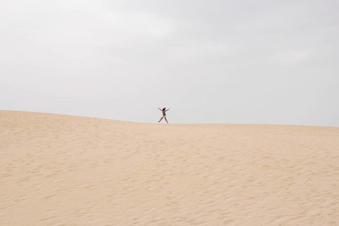 A lone individual joyfully jumps on a vast sand dune under a cloudy summer sky, capturing the