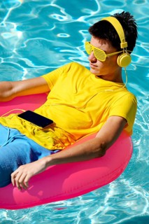 A person in a yellow shirt, sunglasses, and headphones lounges on a pink inflatable float in a