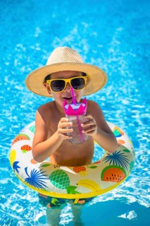 Happy boy wearing sunglasses and hat in the swimming pool drinking water from straw