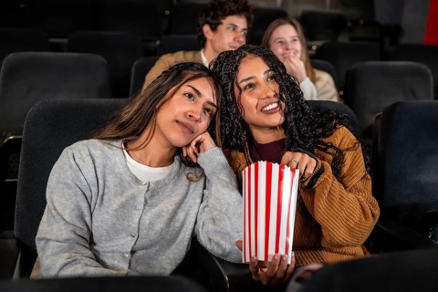 Two friends enjoying a movie at the cinema, one is eating popcorn while the other looks bored