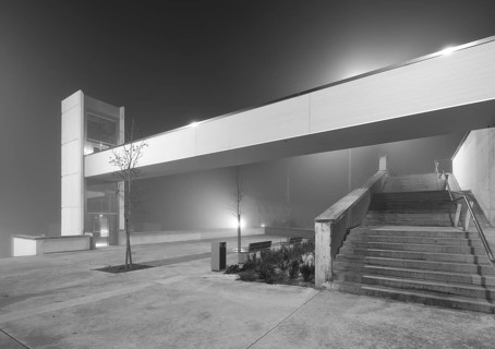 A modern urban elevator and connecting staircase are shrouded in dense mist under a glowing light,