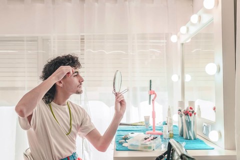 Young man plucking his eyebrows in a bedroom with a vanity mirror surrounded by led lights,