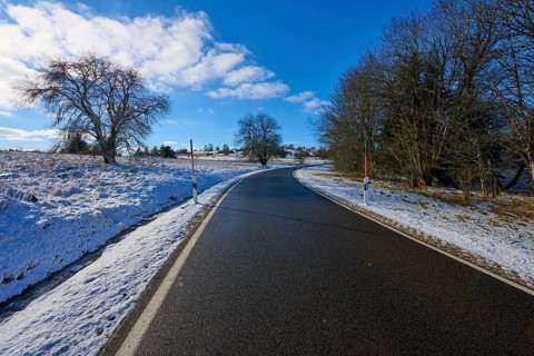 A sunny country road winds through a snow-covered landscape with bare trees and blue sky, winter,