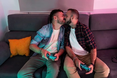 A joyful gay couple shares a kiss while holding gaming controllers, sitting on a comfortable sofa