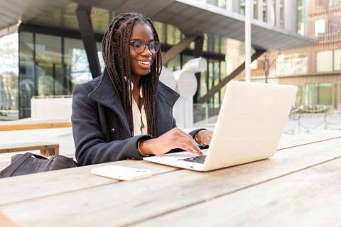 A young African American businesswoman with braids works on a laptop at an outdoor table in a