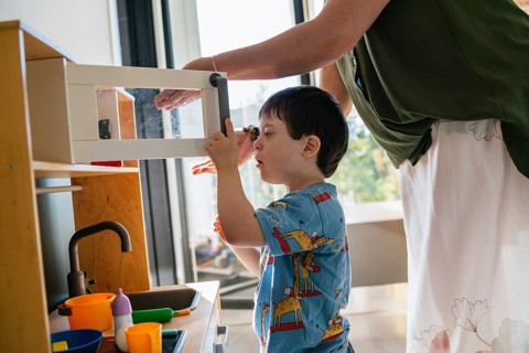 A young child with Down syndrome engages with a kitchen playset under the guidance of an adult.