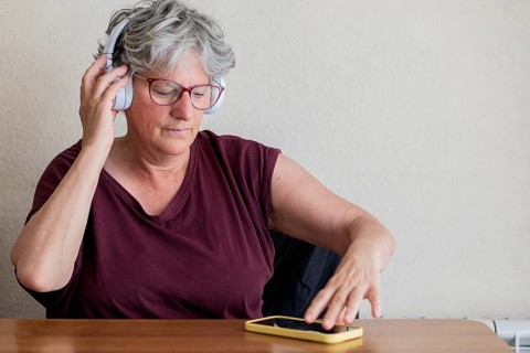 An elderly woman sits at a table, enjoying music through headphones. She uses a smartphone to