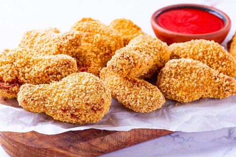 A wooden board holds several golden chicken drumsticks arranged neatly. A small bowl of red dipping