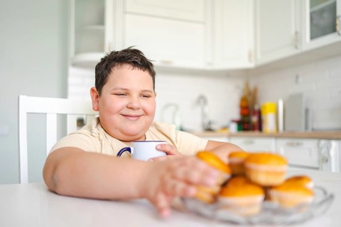 Smiling boy reaches for cupcakes while holding a mug of milk in a bright kitchen. The scene