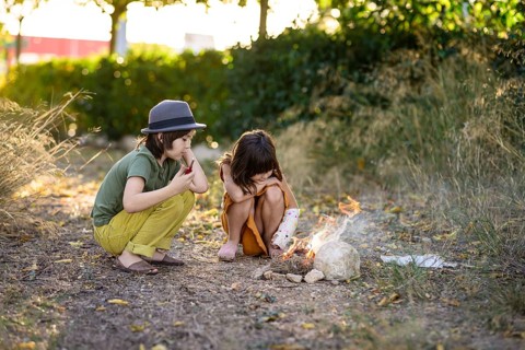 Two children, one in a hat, sit by a small campfire in a natural setting. Surrounded by trees and