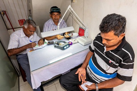 Three men work at a table, one writing while the other examines gemstones under a magnifying glass,