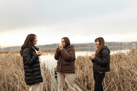 Three women stand by a tranquil lake, engaged in pleasant conversation amidst reeds, each holding a