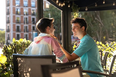 A joyful gay couple sitting at an outdoor cafe, sharing a tender moment. Sunlight filters through
