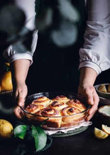 A pair of hands presents a glass dish filled with golden, freshly baked lemon rolls. The rolls are