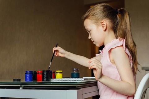 A young girl in a pink dress paints with colorful jars on a table. Engaged and focused, she