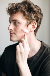 A young male relaxes at home while using a pink quartz facial roller on his skin, focusing on
