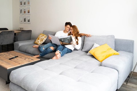 A joyful young couple relaxes on a cozy gray sofa, engaged in reading a large photo album. The warm
