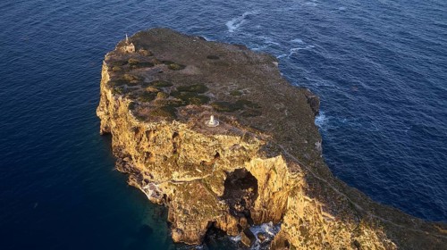Drone shot, rocky island with small lighthouse and cliffs surrounded by calm sea, morning light,