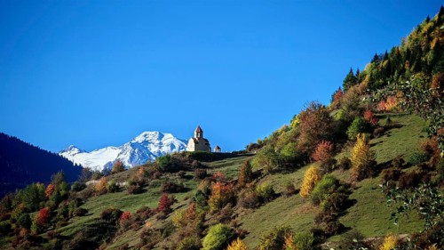 A church on a hill between colorful autumn trees, snow-capped peaks under a blue sky, Mestia,