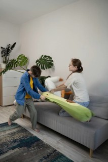 A lively plush toys fight between a young boy and girl on a gray sofa in a cozy living room. The