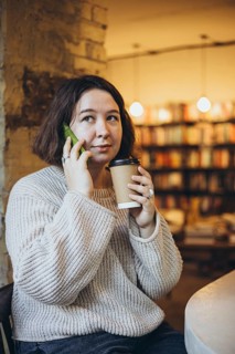 A woman in a relaxed sweater holds a coffee cup and green smartphone while sitting in a bookstore