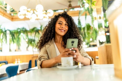 Happy beauty woman smiling at camera while using smartphone sitting in a cafeteria