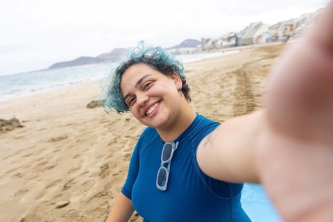 A joyful transgender individual with blue-tinted hair captures a selfie while standing on a sandy