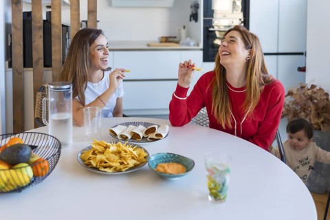 Two cheerful women enjoying tacos and nachos at a table, laughing and sharing a meal, while a baby
