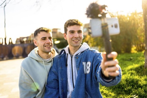 Two men filming a video blog outdoors, using a smartphone attached to a gimbal, smiling and