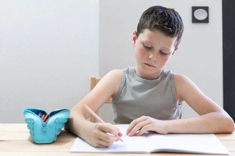 A young student intently writes in a notebook, surrounded by school supplies on a wooden desk. The