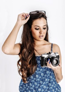 A young woman in a blue patterned dress holds a vintage camera while adjusting her glasses atop her