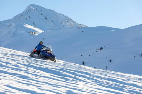 A man rides a snowmobile over a vast, snow-covered mountain slope on a sunny winter day. The snowy