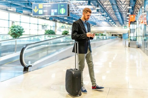 Businessman using smartphone while waiting his flight at the airport terminal, carrying a suitcase