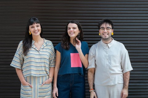 A diverse group of three friends stands together, smiling warmly at the camera. They each wear