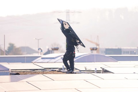 Person on roof lifts solar module surrounded by city and crane, clear daylight scene, PV