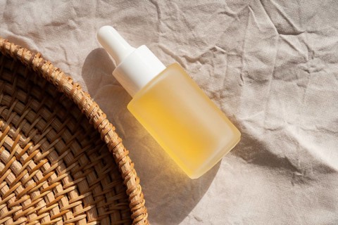 A cosmetic filled with yellow oil rests on textured fabric, next to a woven basket. Sunlit, it