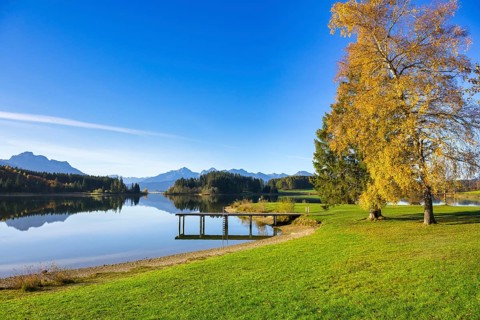 A quiet lake with a jetty, surrounded by autumnal trees and mountains under a clear blue sky,