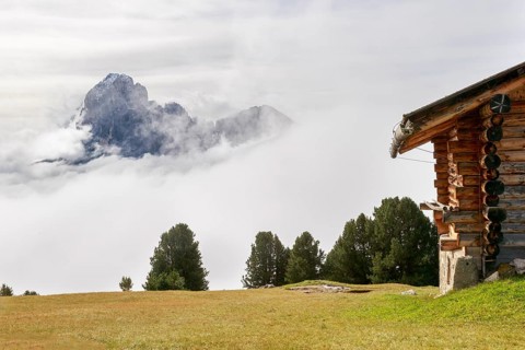 Rustic alpine hut on a green meadow, surrounded by mist and wild mountains in the background