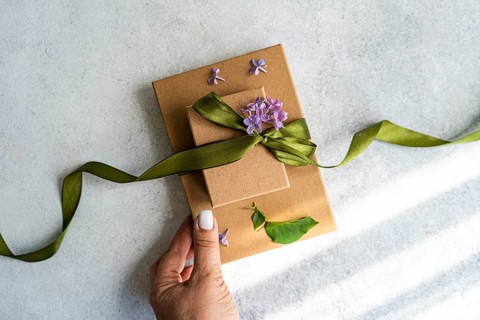 A hand holds a beautifully wrapped gift box adorned with a green ribbon and purple flowers. The